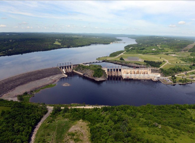 Aerial view of the Mactaquac Generating Station looking towards the arm (North-Northwest). The left side of the dam is earthen, the sluiceway central, and the spillway, turbines, and powerhouse on the right. Mactaquac Road is seen on the top of the embankment and crosses the rest of the river upstream of the turbines.