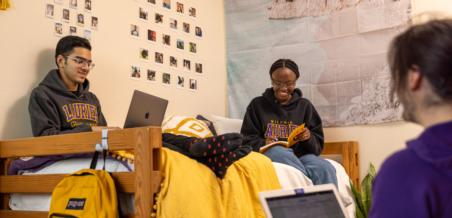 three students in residence room sitting on bed studying